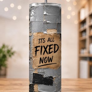 May include: A cylindrical tumbler with a straw featuring a graphic print of overlapping silver and black duct tape strips. A central tan rectangular patch with torn edges displays the black text ITS ALL FIXED NOW in a bold, hand-lettered style.