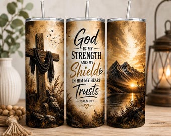 May include: A cylindrical tumbler with a metal straw featuring a sepia-toned design. The graphic includes a wooden cross draped in fabric, a mountain landscape at sunset by a lake, and the text God is my strength and my shield in him my heart trusts Psalm 28:7.