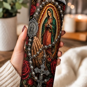 May include: A cylindrical tumbler with a metal straw featuring a religious graphic print of the Virgin of Guadalupe, praying hands, a silver rosary with a crucifix, and deep red roses against a dark, ornate background with golden light rays.