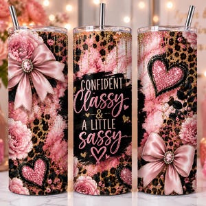 May include: Three cylindrical tumblers with straws featuring a pink and black leopard print, pink floral accents, and glittery heart motifs. The center tumbler displays the text "CONFIDENT Classy & A LITTLE SASSY" in black and pink script lettering.