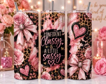May include: Three cylindrical tumblers with straws featuring a pink and black leopard print, pink floral accents, and glittery heart motifs. The center tumbler displays the text "CONFIDENT Classy & A LITTLE SASSY" in black and pink script lettering.