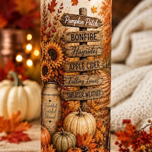 May include: A cylindrical tumbler with a clear lid and metal straw featuring an autumn-themed print. The design includes a wooden signpost with text reading Pumpkin Patch, BONFIRE, Hayrides, APPLE CIDER, Falling Leaves, and SWEATER WEATHER.