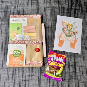 Op de afbeelding: Een cadeaupakket met een leeslijst, thee, een wenskaart met een bloemenontwerp, een pen, plaknotities en een zakje gummy snoepjes. De leeslijst zit in een decoratieve houder met bloemenpatroon. Een rode lieveheersbeestjessticker is ook inbegrepen.