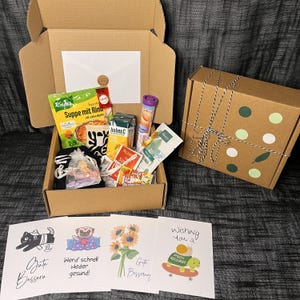 Könnte beinhalten: Eine braune Pappbox gefüllt mit Genesungsgaben. Die Box enthält Suppenpulver, Saft, Tee, Süßigkeiten, ein Stofftier und eine Karte mit der Aufschrift "Wünsche dir gute Besserung".