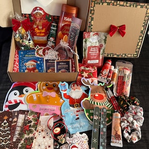 Könnte beinhalten: Eine weihnachtliche Geschenkbox voller verschiedener Artikel. Enthält Gesichtsmasken, Badeprodukte, Lippenbalsam, Make-up-Pinsel und dekorative Aufkleber. Die Box ist mit roten Schleifen und festlichen Verpackungen verziert.