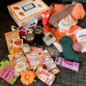 Könnte beinhalten: Eine Auswahl an herbstlichen Artikeln, darunter eine Geschenkbox, ein Schal, eine Handcreme, Gesichtsmasken, Lippenbalsam, Kerzen und eine Schneekugel. Auf der Box steht "My Little Serendipity Box".