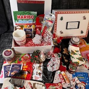 Könnte beinhalten: Eine weihnachtliche Geschenkbox mit einem weißen Becher mit Schneeflockenmuster, einer Tüte Haribo Winter Mix, Meßmer Tee, einem Make-up-Pinselset, einer Duftkerze, festlichen Socken, Haargummis, Lippenbalsam und verschiedenen weihnachtlichen Pflegeprodukten.