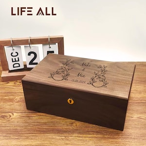 Puede incluir: Una caja de madera con base marrón oscuro y tapa marrón claro. La tapa está grabada con diseños florales y los nombres "Mike & Mia" junto con la fecha "15.06.2024". La caja tiene una cerradura dorada. La marca "LIFE ALL" está en la parte superior.