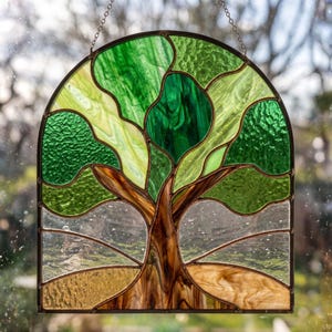 May include: An arched stained glass suncatcher featuring a tree with a brown trunk and branches, surrounded by various shades of green glass leaves. The base consists of textured tan glass, and the piece is suspended by a thin metal chain.