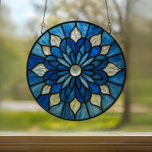May include: A round stained glass window with a floral design. The window features shades of blue and white, with a central clear glass circle. The glass is framed by a black metal border and is suspended by a thin wire.