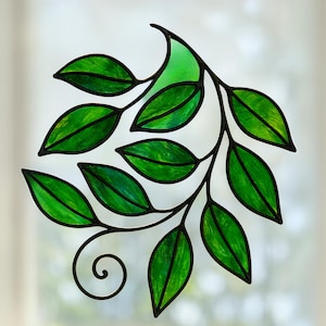 May include: A stained glass leaf design with bright green leaves and black borders. The leaves are arranged on a curved stem with a decorative swirl. The artwork is set against a blurred, light background.