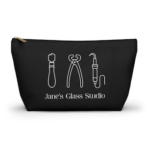 May include: Black zippered pouch featuring white line art of glass working tools: a glass cutter, pliers, and a soldering iron. The text "Jane's Glass Studio" is printed below the tools.