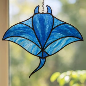 May include: A stained glass manta ray suncatcher. The design features shades of blue with a wavy pattern, outlined in black. It is suspended by a chain and is designed to hang in a window. The measurements are in centimetres.