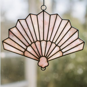 Puede incluir: Un abanico decorativo de vidrieras en tonos rosa y blanco, delineado con líneas negras. Cuenta con un lazo para colgar en la parte superior. El diseño del abanico recuerda a los abanicos tradicionales.