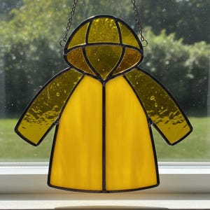 May include: A stained glass art piece depicting a yellow raincoat with a hood. The glass is a translucent yellow with black outlines and is suspended by a chain. The artwork is displayed in front of a window.