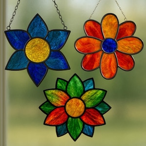 May include: Three stained glass flower suncatchers. One is blue and yellow, another is orange and red, and the third is green, red, and yellow. Each flower is outlined in black and hangs from a chain.