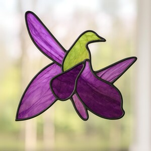 Può includere: Colibrì in vetro colorato con sfumature viola e verdi. Le ali e il corpo dell'uccello sono realizzati in vetro traslucido, bordati con saldatura nera. Il design cattura l'essenza di un colibrì in volo, adatto per la decorazione di finestre.