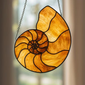 Pode incluir: Apanhador de sol de concha nautilus em vitral com design em espiral. A concha apresenta tons de âmbar e castanho, delineados com metal preto. Uma corrente é anexada para pendurar. Decoração de casa.