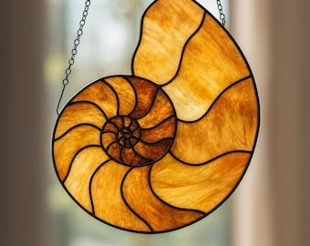May include: Stained glass nautilus shell suncatcher with a spiral design. The shell features shades of amber and brown, outlined with black metal. A chain is attached for hanging. Decorative home decor.