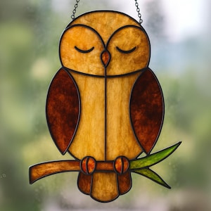 May include: Stained glass owl decoration with a warm color palette of amber, brown, and orange. The owl is perched on a branch with green leaves. The owl has closed eyes, suggesting a peaceful or sleepy state. The piece is suspended by a chain.