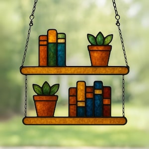 Op de afbeelding: Gebrandschilderd glazen hangende plank met twee niveaus. Elk niveau bevat een terracotta pot met een groene plant en een stapel kleurrijke boeken. De boeken zijn oranje, geel, blauw en groen. De plank wordt opgehangen door een zwarte ketting.