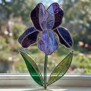 Może przedstawiać: Witrazowy kwiat irysa w odcieniach fioletu, niebieskiego i zieleni. Kwiat ma szczegółowy wzór z łodygą i dwoma liśćmi. Szkło jest oprawione w metal w kolorze srebrnym.