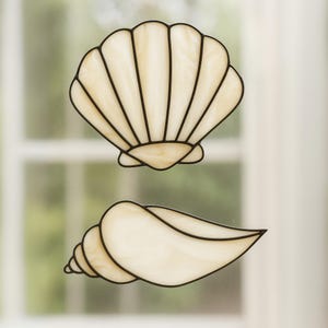 Puede incluir: Dos decoraciones de conchas marinas de vidrieras. La superior es una concha de vieira en forma de abanico, y la inferior es una concha en espiral. Ambas son de color crema con contornos negros, diseñadas para colgar en una ventana.