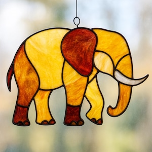 Könnte beinhalten: Glasmalerei-Elefant mit schwarzer Umrandung. Der Elefant ist in Gelb-, Orange- und Brauntönen gehalten. Das Ohr des Elefanten ist tiefrotbraun und die Stoßzähne sind weiß. Der Elefant hängt an einem schwarzen Draht.