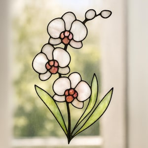 Puede incluir: Una orquídea de vidriera con pétalos blancos, centros rosas y hojas verdes. La flor tiene un contorno negro y se encuentra sobre un fondo borroso. La orquídea es una pieza decorativa.