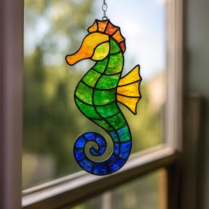 May include: A colorful stained glass seahorse suncatcher. The seahorse features a gradient of colors, including yellow, orange, green, and blue. The seahorse is hanging in front of a window, with a blurred background of green foliage.