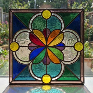 May include: Square stained glass panel with a geometric floral design. The panel features a central flower with colorful petals in red, orange, yellow, green, and purple. The background is composed of blue, green, and clear glass sections, with yellow circles at the corners.