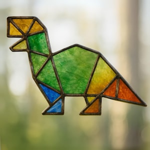 May include: A stained glass dinosaur suncatcher in a geometric design. The dinosaur features a variety of colors, including yellow, green, blue, and orange, with a dark metal frame. The suncatcher is designed to be hung in a window.
