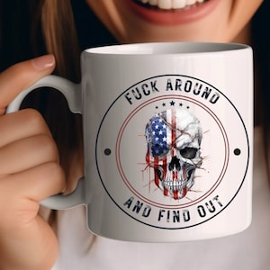 May include: White ceramic coffee mug with a black and white graphic of a skull with an American flag design. The text "F**K AROUND AND FIND OUT" is printed in black around the skull.