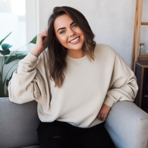 Peut inclure: Une femme aux longs cheveux bruns sourit et porte un sweat-shirt beige clair et un pantalon noir. Elle est assise sur un canapé gris.