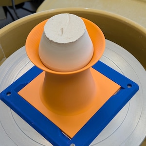 May include: A pottery wheel with a white clay form in the center. The clay sits on an orange and blue support structure. The wheel is yellow and the background is blurred.