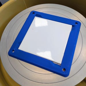 May include: A square white ceramic tile centered within a blue plastic frame with four circular corner holes, positioned flat on a circular metallic pottery wheel surface that is set inside a yellow basin with a visible power switch.