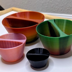 May include: Four divided bowls in different sizes and colors. The largest bowl is orange and red, the next is green and orange, the third is pink and orange, and the smallest is black.