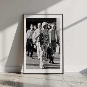 May include: Black and white framed print of an astronaut in a spacesuit walking with a group of men. The astronaut is the focal point, wearing a helmet and a detailed spacesuit. The image is a historical photograph.