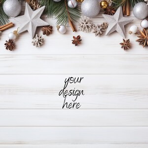 Pu&ograve; includere: Vista dall'alto di una superficie in legno bianco decorata con ornamenti natalizi. Palline argentate e dorate, decorazioni a forma di stella, bastoncini di cannella e rami di pino sono disposti in alto. Il testo "your design here" &egrave; scritto in corsivo.