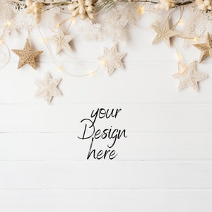 Pu&ograve; includere: Una scena festiva natalizia con ornamenti a stella dorati e bianchi, fiocchi di neve e luci a catena su uno sfondo di legno bianco. Il testo "your Design here" &egrave; scritto in corsivo.