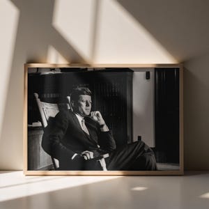 May include: Black and white framed photograph of a man in a suit seated in a wooden rocking chair. The photograph is in a light-coloured wooden frame and is displayed against a neutral wall.