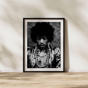 May include: Monochrome framed print featuring a man with a large afro, adorned with a patterned jacket and necklaces. The portrait is a medium shot, with the subject looking downwards. The frame is black, set against a neutral backdrop.
