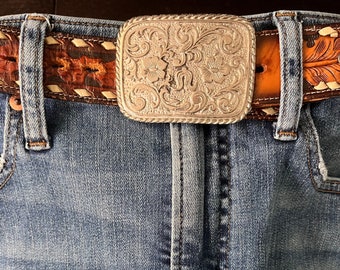 Puede incluir: Una hebilla de cinturón plateada con un diseño floral, puesta en un par de pantalones vaqueros azules con un cinturón de cuero marrón.