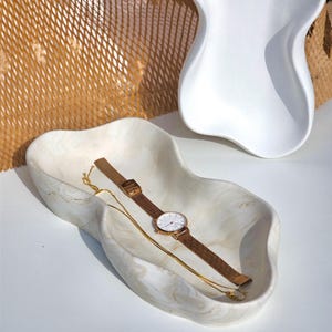 May include: A white marble-like tray with a gold watch and a gold chain necklace on top. The tray has an irregular, organic shape.