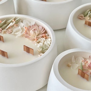 May include: Five white ceramic candle holders with a wide, rounded base. Each holder contains a white candle with dried flowers and greenery embedded in the wax. Wooden wicks are visible in some of the candles.