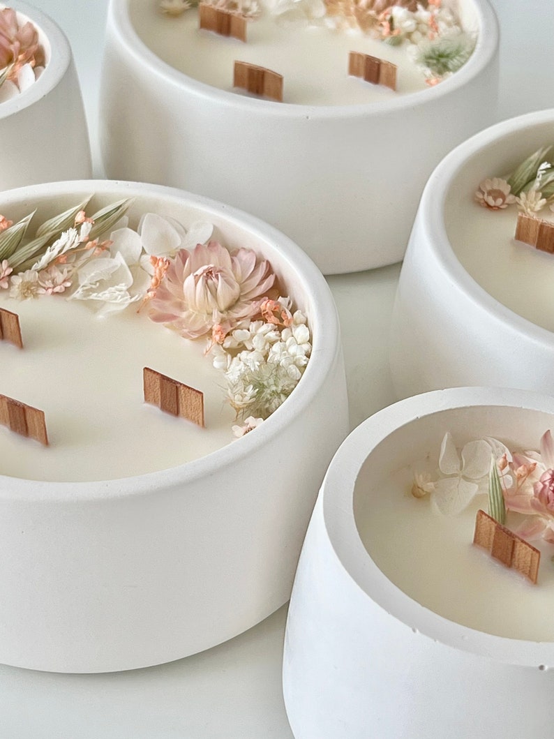 May include: Five white ceramic candle holders with a wide, rounded base. Each holder contains a white candle with dried flowers and greenery embedded in the wax. Wooden wicks are visible in some of the candles.