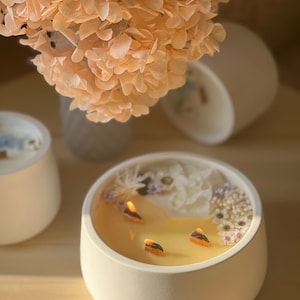 May include: A white ceramic candle holder with a yellow candle and dried white flowers on top. The candle has three wicks and is lit.