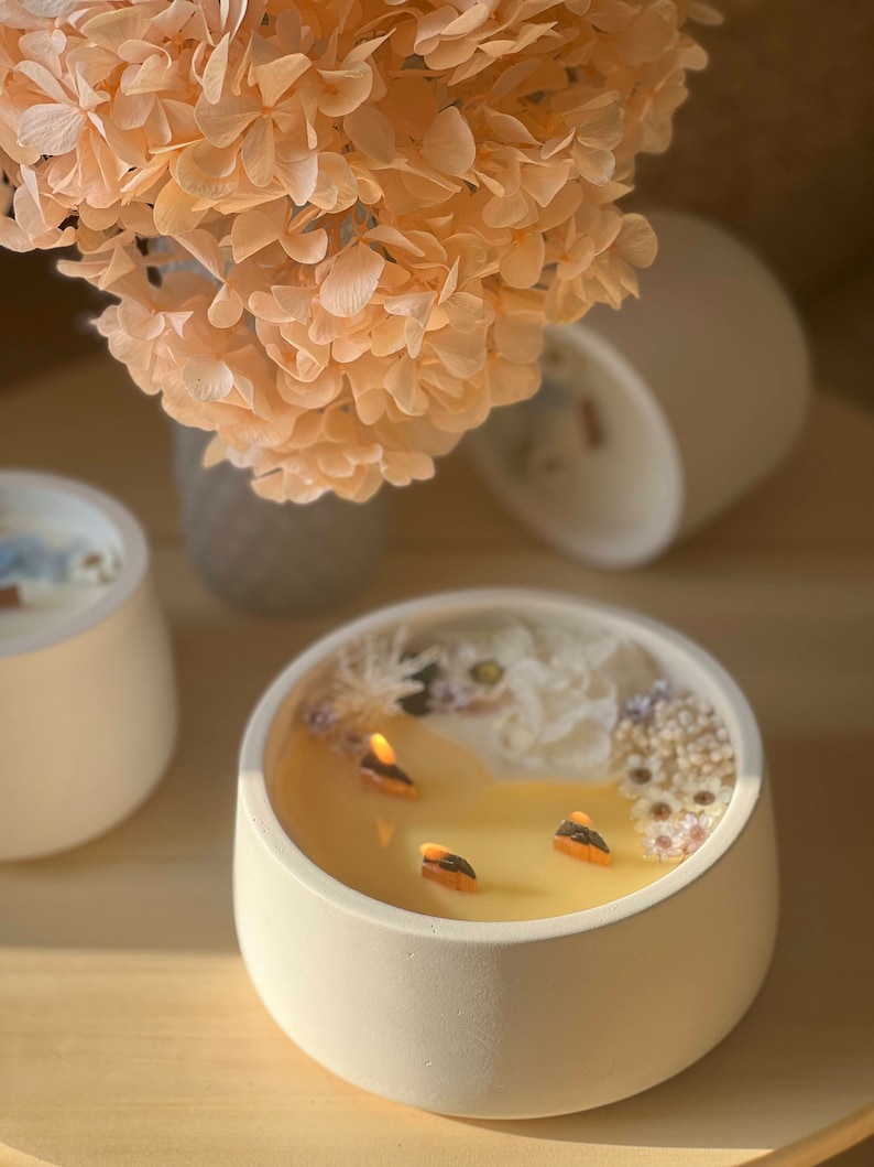 May include: A white ceramic candle holder with a yellow candle and dried white flowers on top. The candle has three wicks and is lit.