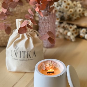 May include: A white ceramic candle jar with a lid. The jar is filled with a white candle and has dried flowers embedded in the wax. The candle is lit and the flame is visible. The jar is sitting on a wooden surface. The text "CVITKA natural candles & flowers" is printed on a white fabric bag in the background.