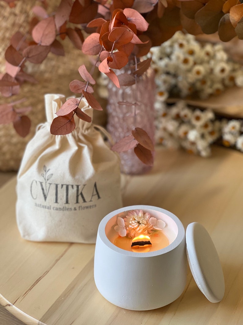 May include: A white ceramic candle jar with a lid. The jar is filled with a white candle and has dried flowers embedded in the wax. The candle is lit and the flame is visible. The jar is sitting on a wooden surface. The text "CVITKA natural candles & flowers" is printed on a white fabric bag in the background.